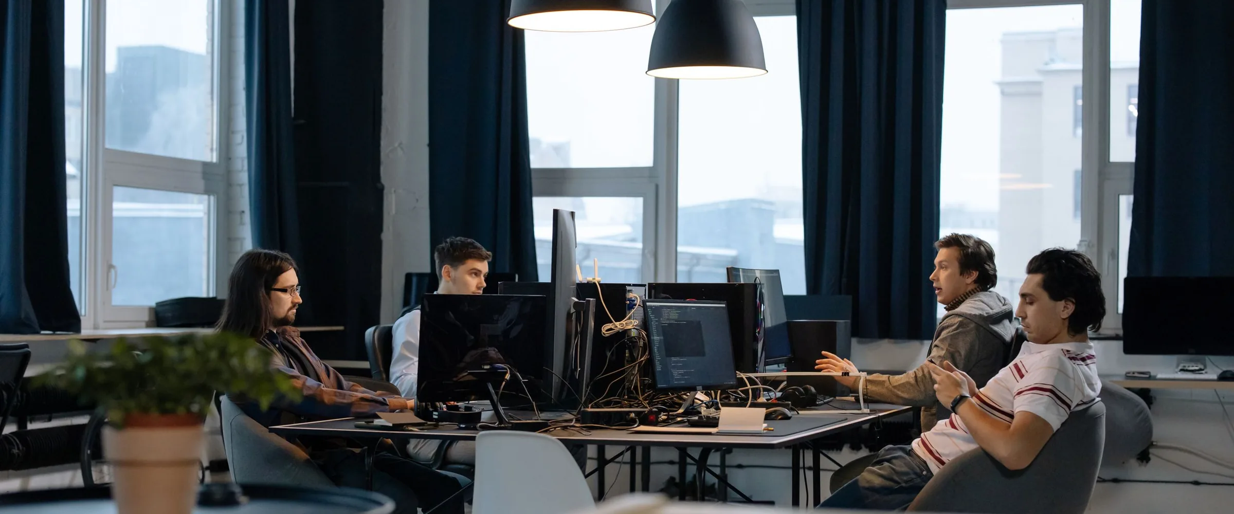 A software team at their desks under hanging lamps, one person talking across the table.
